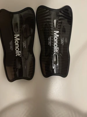Monolit benskydd i kolfiber - Säljer ett par svarta benskydd från Monolit, tillverkade i kolfiber med ergonomisk form för bästa passform. Lätta och slitstarka, perfekta för fotboll eller andra sporter där skydd behövs. Snygg design med vit logga på framsidan.