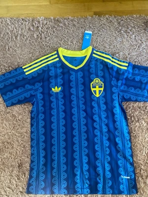 Blå Sverige fotbollströja Adidas - Säljer en blå fotbollströja från Adidas med Sveriges landslagsemblem på bröstet. Tröjan har gula detaljer, tre gula ränder på axlarna och ett mönster i olika blå nyanser. Tillverkad i ett lätt och ventilerande material, perfekt för träning eller match.