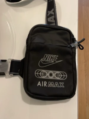 Nike Air Max svart shoppingväska - Snygg svart shoppingväska från Nike Air Max med justerbar axelrem och coolt tryck framtill. Väskan har dragkedja, mesh på baksidan och detaljer i grått. Perfekt för att bära det viktigaste och matcha till streetwear-stilen.