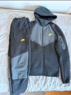 Nike grå och svart loungewear set - Snyggt loungewear set från Nike i mörkgrå och ljusgrå färg med gula Nike-loggor. Setet består av en hoodie med dragkedja, huva och fickor samt matchande byxor med dragsko i midjan. Perfekt för chill dagar hemma eller när du vill vara bekväm.