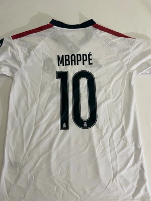 Real Madrid Mbappé matchtröja - Snygg vit Real Madrid matchtröja med Mbappé och nummer 10 på ryggen. Tröjan har svarta detaljer, röda axelränder och klubbmärke på bröstet. Tillverkad i lätt och ventilerande material, perfekt för fotboll eller träning. Adidas-logga och sponsortryck på framsidan.