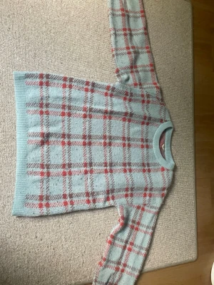 Ljusblå rutig stickad tröja - Säljer en ljusblå stickad tröja med rött och grått rutmönster. Tröjan har rund halsringning och ribbade muddar vid ärmslut och nederkant. Perfekt för dig som gillar en mysig och trendig look med retrovibbar.