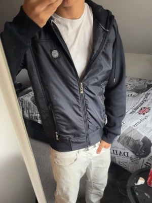 Svart cardigan från Stone Island - Säljer en svart hoodie från Stone Island med dragkedja framtill och två fickor. Ikonisk Stone Island-patch på vänster ärm och logga på bröstet. Hoodien har ribbade muddar och en skön huva. Materialet är en mix av bomull och nylon för en modern look. Pris kan diskuteras!