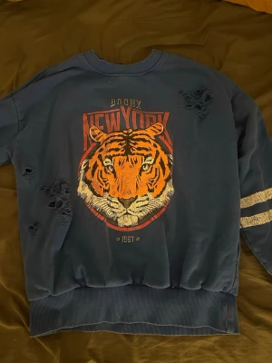 Mörkblå sweatshirt med tigertryck - Cool mörkblå sweatshirt med stort tigertryck och texten 'Bronx New York 1987' på bröstet. Tröjan har slitna detaljer och två vita ränder på ena ärmen. Tillverkad i mjukt bomullsmaterial och har en avslappnad passform. Perfekt för dig som gillar streetstyle.