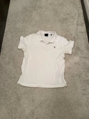 Klassisk vit GANT Pikétröja / Polo (Stl M) - Stilren och fräsch vit pikétröja från GANT med den klassiska broderade loggan på bröstet. Perfekt basplagg till garderoben! 