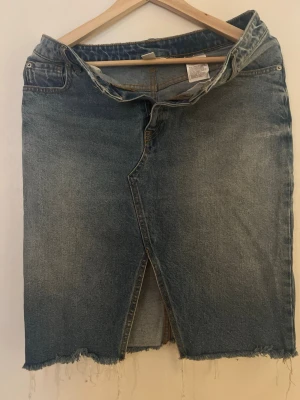 Blå jeanskjol med rå kant från H&M - Säljer en blå jeanskjol från H&M i storlek 40. Kjolen har en rak passform, rå kant nertill och en snygg slits framtill. Klassisk femficksmodell med dragkedja och knapp. Perfekt för en avslappnad och trendig look.