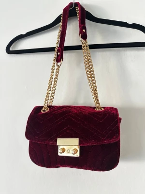 Vinröd sammetsväska med guldkedja - Snygg vinröd handväska i mjuk sammet med quiltat mönster. Väskan har en guldfärgad kedjerem och ett elegant guldfärgat lås framtill. Perfekt accessoar för att lyfta din outfit med en lyxig touch.