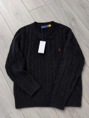 Svart kabelstickad tröja Polo Ralph Lauren - Kabelstickad svart tröja från Polo Ralph Lauren med rund halsringning och röd broderad logga på bröstet. Tröjan har ribbade muddar vid ärmslut och nederkant. Perfekt för dig som gillar klassisk och stilren design.