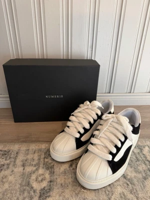 Svarta och vita sneakers från Numeris - Snygga sneakers i svart och vitt från Numeris. Skorna har breda vita snören, vit tå med räfflad struktur och svart mockadetalj längs sidan. Ovandelen är i textil och mocka, sulan är vit och tjock för en modern look.