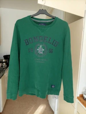 Grön sweatshirt från Bondelid - Grön sweatshirt från Bondelid med tryck framtill och ribbade muddar vid ärmslut och nederkant. Klassisk rund halsringning och avslappnad passform. Perfekt för en chill och stilren look. Finns en liten fläck ja int först få bort 