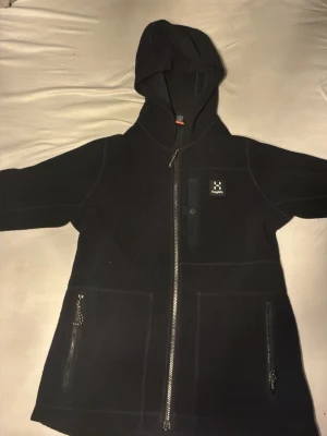 Svart fleecehoodie från Haglöfs - Mysig svart hoodie i fleece från Haglöfs med dragkedja framtill, två stora fickor med dragkedja och en mindre bröstficka. Klassisk huva och diskret logga på bröstet. Perfekt för lager-på-lager och chill dagar. Passar perfekt för hösten och vintern.