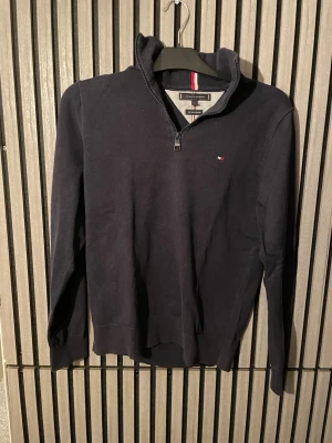 Mörkblå halvzip-tröja Tommy Hilfiger - Mörkblå tröja med halvzip och ståkrage från Tommy Hilfiger. Diskret logga på bröstet och ribbade muddar vid ärmslut och nederkant. Perfekt för lager-på-lager och snygg till jeans. Materialet är mjukt och bekvämt.