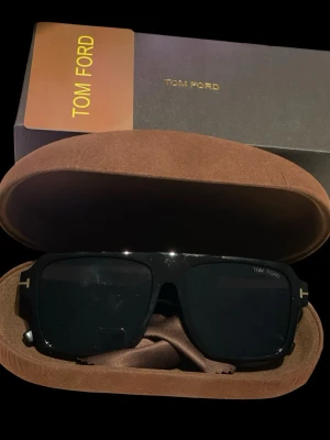 Svarta solglasögon från Tom Ford - Snygga svarta solglasögon från Tom Ford med rektangulär form och mörka glas. Kommer med originalfodral och box, perfekt för dig som vill ha en stilren accessoar med exklusiv känsla. Klassisk Tom Ford-design med diskret logga på glaset. Sparsamt använda under sommaren. Säljes prutat och klart till fyndpris / ”höstpris”. 