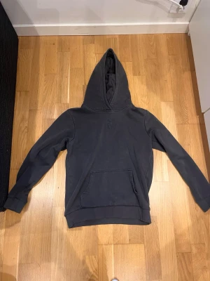 Mörkgrå hoodie från peak performance  - Säljer en mörkgrå hoodie från peak performance med stor huva och magficka. På ryggen finns ett svart geometriskt tryck som ger en cool touch. Hoodien har ribbade muddar vid ärmar och nederkant. Perfekt för dig som gillar enkel men stilren streetwear.