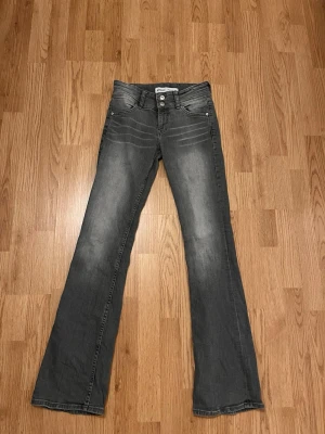 Grå bootcut jeans med dubbla knappar - Snygga grå jeans med bootcut-modell och dubbla knappar i midjan. Jeansen har klassiska fem fickor och diskreta slitningar framtill för en cool look. Perfekta till sneakers eller boots för en avslappnad stil.