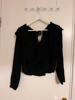Svart blus med volanger och knytning - Trendig svart blus med långa ärmar, volangdetaljer längs bröstet och axlarna samt knytning i nacken. Blusen har en lätt croppad passform och dekorativa spetsdetaljer vid halsringningen. Perfekt för en stilren och chic look.