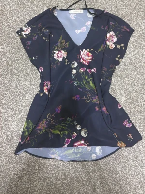 Mörkblå blommig 2000s vintage shirt - Supersnygg mörkblå kortklänning 2000 stil vintage med blommigt mönster i rosa, vitt och grönt. Klänningen har v-ringning och korta ärmar, perfekt för sommarens alla tillfällen. Materialet känns lätt och svalt mot huden.