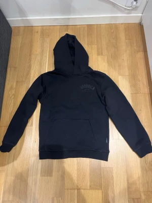 Svart hoodie från Hoodrich - Svart hoodie från Hoodrich med stor huva och magficka. Tryckt Hoodrich-logga på bröstet och större tryck på ryggen. Klassisk passform och ribbade muddar vid ärmslut och nederkant. Perfekt för en avslappnad streetwear-look.