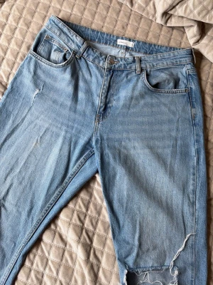 Ljusblå slitna bootcut jeans - Säljer ett par ljusblå jeans med raka ben och slitningar på låret och knät. Jeansen har klassisk femficksmodell, bälteshällor och knappgylf. Perfekta för en avslappnad och trendig look. Dem är tyvärr för stora på mig därför säljer jag dem. 