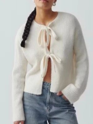 Vit cardigan med snörning - Trendig vit ribbstickad kofta med tre knytband framtill. Koftan har rund halsringning och långa ärmar. Perfekt att styla över ett linne eller t-shirt för en avslappnad look. Materialet ser mjukt och lite fluffigt ut.