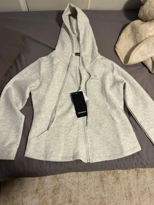 Ljusgrå zip hoodie från luluemon - Ljusgrå hoodie med dragkedja från Sail Racing. Klassisk modell med huva och dragsko, två fickor framtill och diskret logga på bröstet. Tillverkad i mjukt material som passar perfekt till chill eller streetwear. Enkel och stilren design.