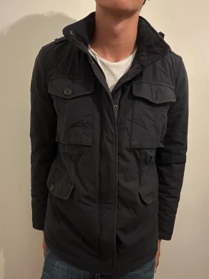Mörkblå field jacket med fickor - Stilren mörkblå skepparkavaj med fyra stora fickor framtill och knappar. Jackan har hög krage och dragkedja samt knappstängning. Materialet ser ut att vara bomullsmix med en matt finish. Perfekt för dig som vill ha en klassisk och funktionell look. Saknar en knapp