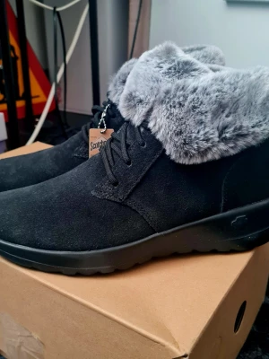 Svarta boots med fuskpäls från Skechers stl 37 - Snygga svarta boots från Skechers, stl 37, med mjuk grå fuskpäls runt skaftet och snörning framtill. Yttermaterialet är mockaliknande och sulan är i svart gummi. Perfekta för kalla dagar när du vill hålla stilen och värmen. Helt nya och oanvända i kartong!