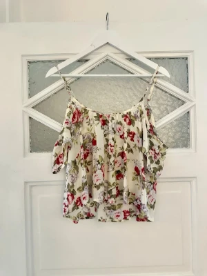 Blommig topp från Abercrombie & Fitch - Supersöt linnetopp från Abercrombie & Fitch med blommigt mönster i rött, grönt och lila på vit botten. Toppen har tunna axelband och en luftig, flowy passform som känns perfekt för varma dagar.
