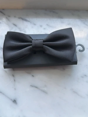 Svart klassisk fluga - Snygg svart fluga i klassisk modell, perfekt för att lyfta din outfit till nästa nivå. Flugan är tillverkad i ett slätt tyg med diskret glans och har en stilren, tidlös form. Passar till skjorta och kavaj för en elegant look.