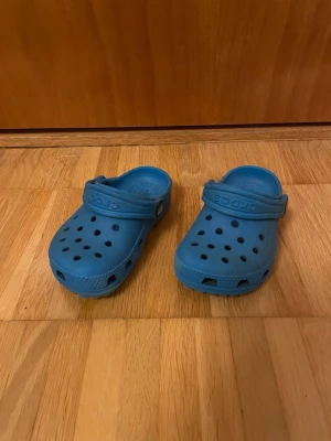Blå Crocs tofflor med rem - Säljer ett par klassiska blå Crocs tofflor med justerbar hälrem och ventilerande hål på ovansidan. Tillverkade i lätt och bekvämt gummimaterial som är perfekt för både inomhus och utomhus. Enkla att rengöra och superpraktiska för vardagen. Är lite smutsiga men är nog lätt att tvätta av dom lite. 