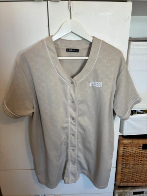 Beige kortärmad skjorta FB Sister - Snygg beige skjorta från FB Sister med korta ärmar och vita detaljer. Skjortan har tryckknappar framtill, vit piping längs kanterna och diskret mönster i tyget. På bröstet står det 'FSHN' broderat i vitt.