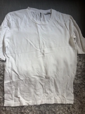 Vit stickad t-shirt med rund hals - En basic vit stickad t-shirt med rund halsringning och halvlånga ärmar. T-shirten har en enkel design och knappdetalj bak i nacken. Tillverkad i mjukt tyg som känns skönt mot huden. Storlek är L men passar mig som brukar ha S.
