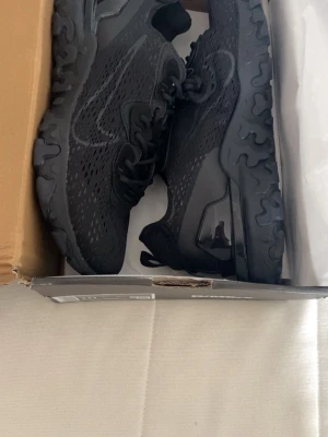 Svarta Nike sneakers med snörning/ Nike react vision - Snygga helsvarta sneakers från Nike med ventilerande mesh-överdel och robust sula. Skorna har klassisk snörning och en modern, sportig design med diskret Nike-logga på sidan. Perfekta för dig som gillar stilrena och bekväma sneakers.