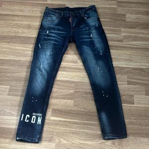Dsquared2 ICON jeans blå  - Snygga blå jeans från Dsquared2 med ikonisk 'ICON'-print på benet och coola slitningar samt färgstänk. Klassisk femficksmodell med smal passform och tydliga tvättade detaljer. Perfekta för dig som vill sticka ut med streetstyle-vibe.