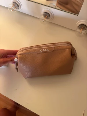Beige necessär från CAIA - Snygg beige necessär från CAIA i ett stilrent och mjukt konstläder. Praktisk dragkedja upptill och rymlig form som rymmer allt du behöver till smink eller hudvård. Perfekt att ha med sig i väskan eller på resan.