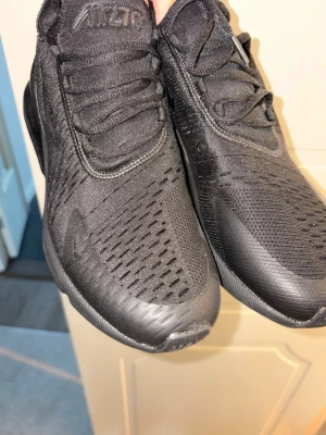 Svarta Nike Air Max 270 sneakers - !HELT NY!Snygga helsvarta Nike Air Max 270 sneakers med mesh-ovandel och tydliga Air Max-detaljer. Skorna har en sportig look med tjock sula och diskret Nike-logga på sidan. Perfekta för dig som gillar streetstyle och vill ha bekväma sneakers med modern design.