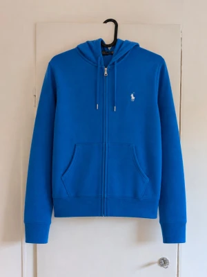 Polo Ralph Lauren zip-hoodie i riktigt snygg klarblå färg 💙 - Klassisk modell med hel dragkedja, huva med dragsko, två framfickor och den ikoniska vita broderade Polo-loggan på bröstet. Storlek M. Snygg och tidlös hoodie som passar perfekt till vardags. Fint begagnat skick utan defekter! 🏷️ Polo Ralph Lauren 📏 Storlek M 🎨 Klarblå med vit broderad logga