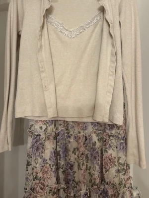 Blommig klänning med spetsdetalj - Romantisk klänning med blommigt mönster i lila, rosa och grönt. Överdelen är ljusbeige med långärmad topp och spetskant vid urringningen. Kjolen är volangig och har ett skirt tyg med blommor. Perfekt för vår och sommar.