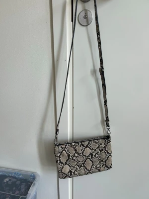 Axelväska med ormmönster - Snygg axelväska i beige och svart med trendigt ormmönster. Väskan har en justerbar axelrem och dragkedja upptill. Perfekt storlek för det viktigaste och ger en cool touch till din outfit. Materialet ser ut att vara konstläder.