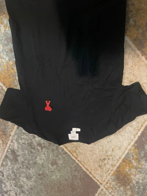 Svart t-shirt med rött hjärtdetalj Ami - Snygg svart t-shirt från Ami med klassisk passform och rund hals. På bröstet sitter Ami:s ikoniska röda hjärtdetalj. Tillverkad i mjuk bomull som känns skön mot huden. Perfekt för dig som gillar stilrena plagg med en twist.