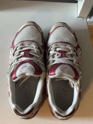 Vita och vinröda Asics sneakers - Säljer ett par vita och vinröda Asics sneakers med mesh- och syntetmaterial. Skorna har klassisk Asics-logga på sidan och detaljer i vinrött och grått. Lågt skaft och snörning framtill. Perfekta för dig som gillar sportig stil.