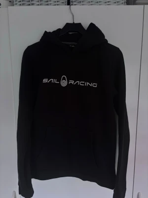 Svart hoodie från Sail Racing - Svart hoodie från Sail Racing med vit logga tryckt på bröstet och en mindre logga på huvan. Klassisk känguruficka framtill och justerbar huva. Perfekt för en avslappnad och sportig stil. Tillverkad i mjukt material för extra komfort.