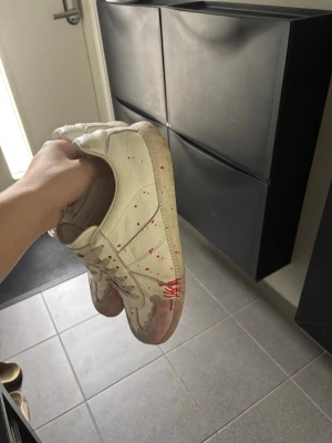 Vita låga sneakers med färgstänk - Säljer ett par vita sneakers med lågt skaft och coola röda färgstänk. Skorna har klassisk snörning och är gjorda i slätt läder. Perfekta för dig som gillar en unik och kreativ stil.