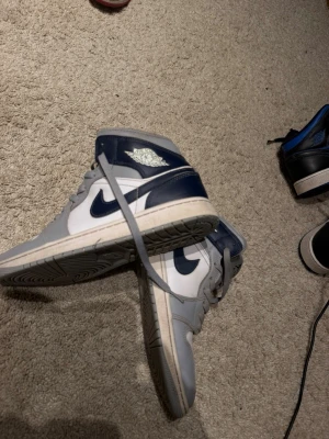 Nike Air Jordan 1 Mid Grå/Marinblå - Snygga Nike Air Jordan 1 Mid sneakers i grått och marinblått med klassisk swoosh på sidan. Skorna har vita paneler, grå snörning och en hög siluett. Tillverkade i läder med perforerad tåbox för extra ventilation. Perfekta för dig som gillar streetwear.