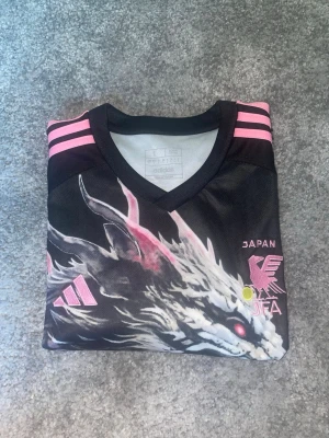 Japan Adidas tröja med drakmotiv - Säljer en svart och rosa sporttröja från Adidas med Japan-tema. Tröjan har ett stort drakmotiv på framsidan, rosa detaljer och tre ränder på axlarna. Tryck med körsbärsblommor och japansk pagod ger en unik look. Perfekt för dig som vill sticka ut på planen.Priset kan diskuteras 