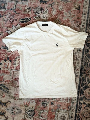 Vit t-shirt från Polo Ralph Lauren - Klassisk vit t-shirt från Polo Ralph Lauren med den ikoniska lilla loggan broderad på bröstet. T-shirten har rund halsringning och korta ärmar. Tillverkad i mjuk bomull för en skön och enkel stil.