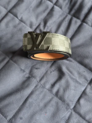 Rutigt Louis Vuitton bälte i skinn - Snyggt bälte från Louis Vuitton med klassiskt rutigt mönster i grått och beige. Bältet har en stor metallspänne med LV-logga och är gjort i skinn. Perfekt accessoar för att lyfta din outfit.