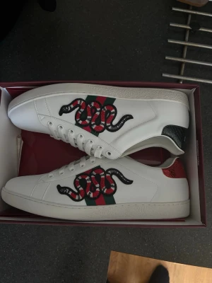 Gucci vita sneakers med orm - Säljer ett par vita Gucci sneakers med ikonisk orm-brodyr på sidan och gröna/röda detaljer. Skorna har lågt skaft och klassisk snörning. Hälpartiet har metallicröda och gröna detaljer för extra stilpoäng. Perfekta för dig som vill sticka ut.
