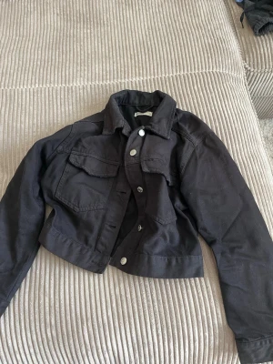 Svart croppad jeansjacka - Snygg svart croppad jeansjacka med klassisk krage och silverfärgade knappar. Jackan har två stora bröstfickor med lock och raka ärmar. Perfekt för dig som vill ha en trendig och enkel look.