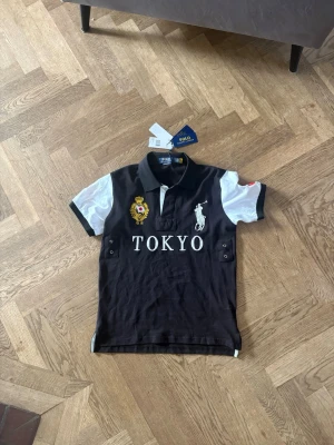 Japan cheif keef - Säljer en svart och vit pikétröja från Polo Ralph Lauren med TOKYO-brodyr, guldfärgad patch och stor logga på bröstet. Klassisk krage och korta ärmar. Perfekt för dig som gillar sportig och stilren look.
